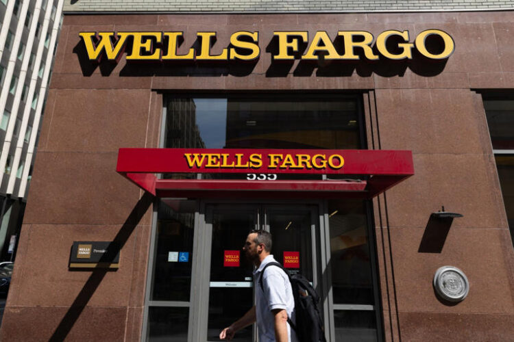 A man in a white shirt walks in front of a Wells Fargo branch with the Wells Fargo logo displayed above the doors.